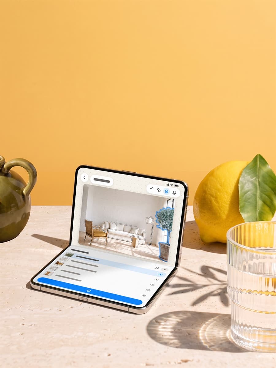 A half-open foldable phone on a stone table in bright sun, showing a living room split into layers with its olive tree selected