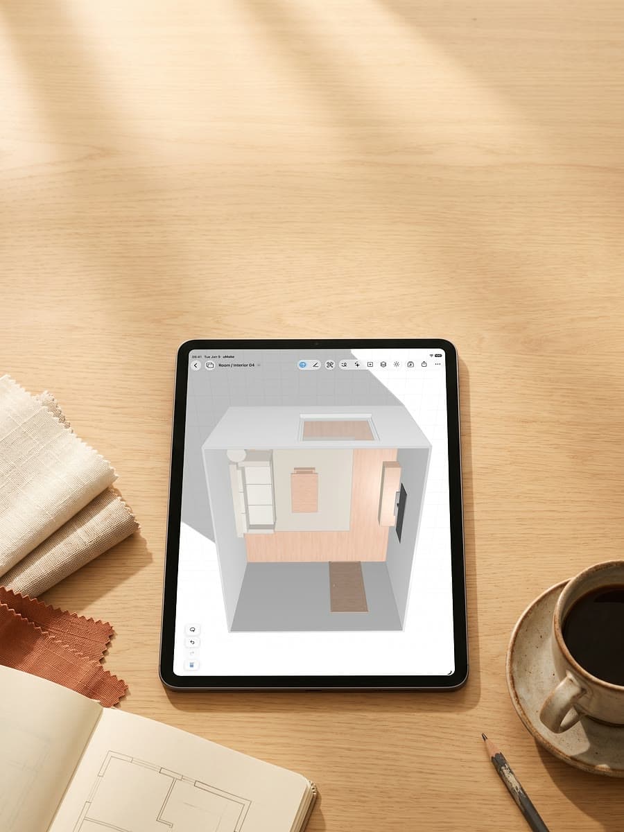 An iPad on a sunlit wooden desk showing a 3D room from above, beside fabric swatches, a cup of coffee and a sketchbook with a floor plan