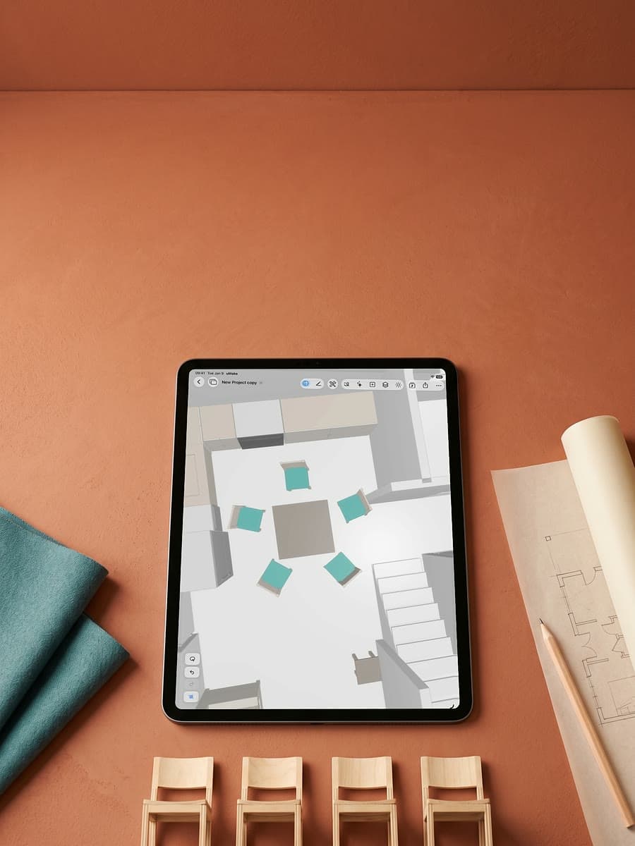 An iPad on a terracotta desk showing a room from above with five matching teal chairs around a table, beside a row of wooden model chairs and rolled plans